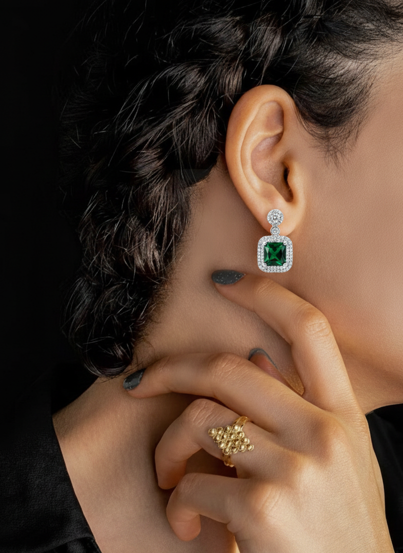 Close-up of a woman wearing square green gemstone and diamond halo earrings and a gold textured ring