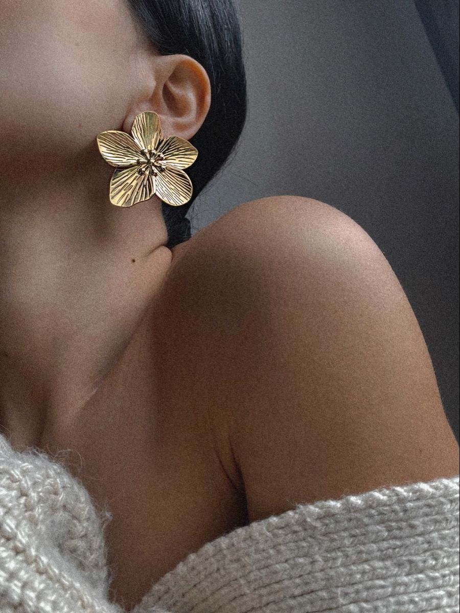 Close-up of a woman's ear wearing a large gold flower-shaped earring and a white knitted sweater